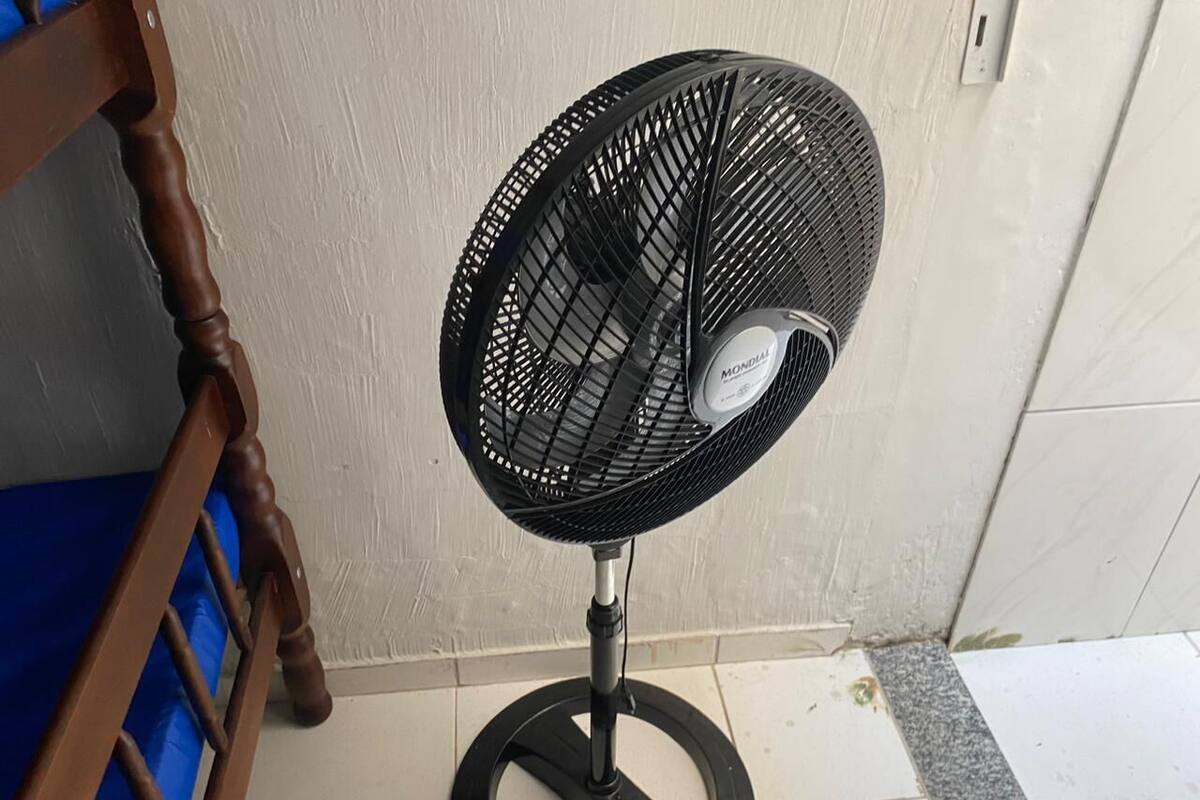 Un ventilateur sur pied, doté d’une grille circulaire et d’un socle, placé contre un mur, au-dessus d’un sol carrelé.