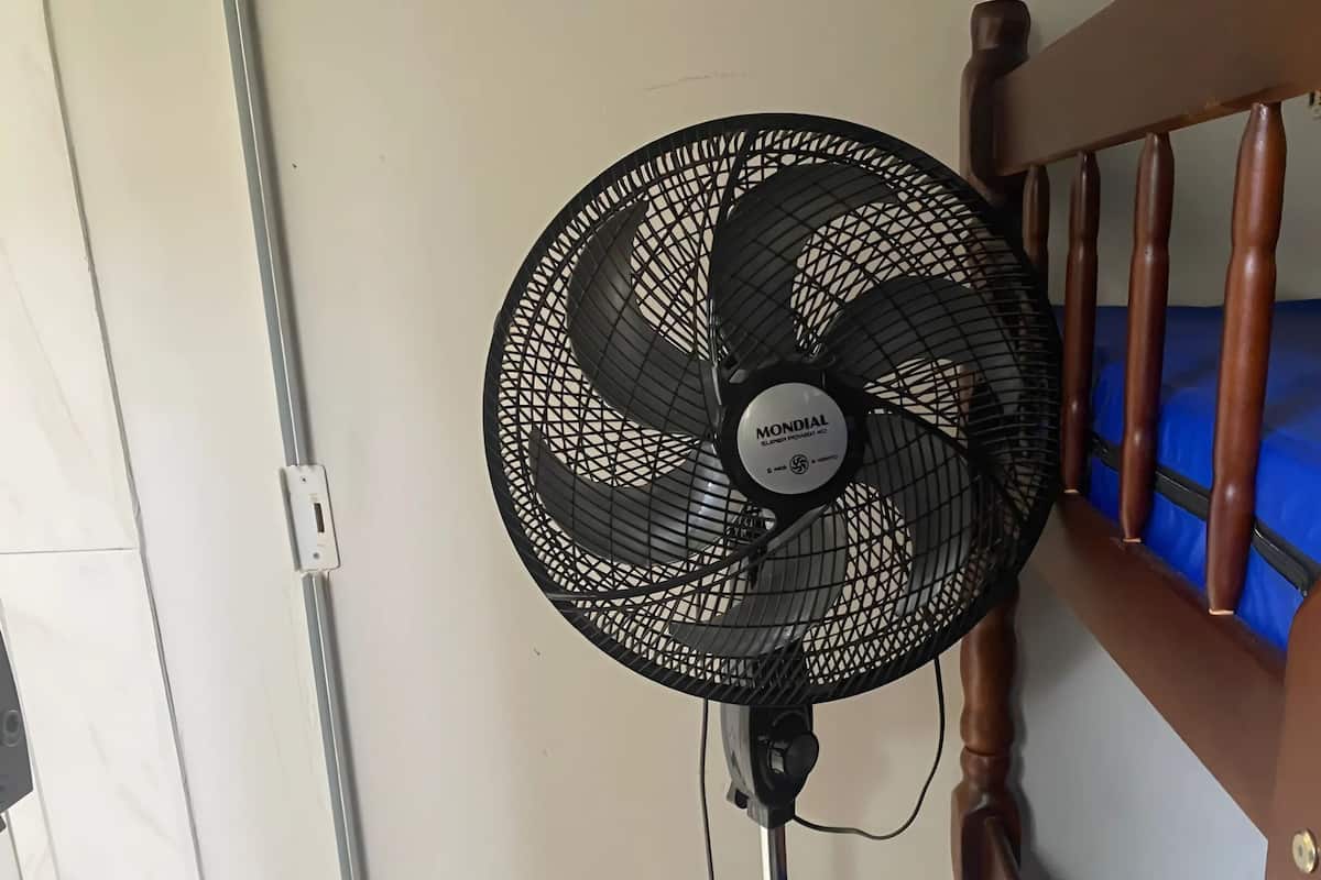 Un ventilateur noir, muni d’une étiquette, est fixé au mur, à côté du cadre en bois d’un lit.