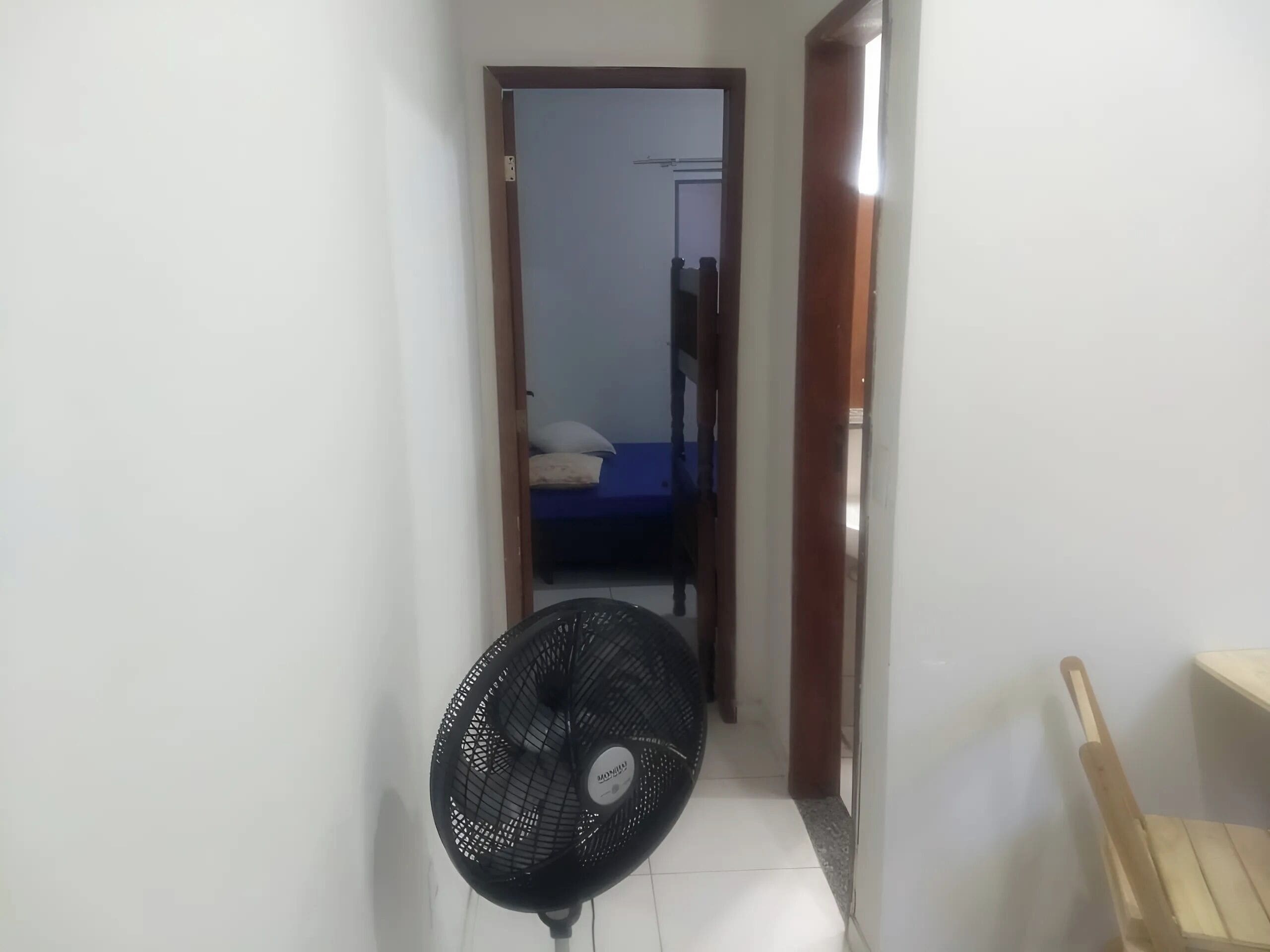 Un couloir étroit, dans lequel on aperçoit un ventilateur noir, une chaise en bois et une porte donnant sur une pièce où se trouve un lit avec un matelas bleu.