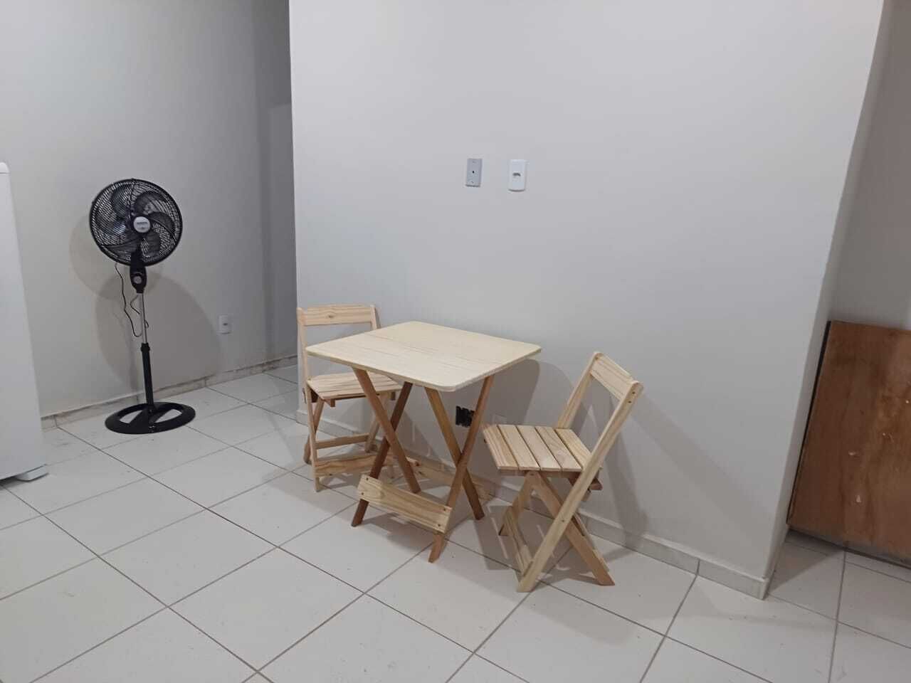 A small room with a white tiled floor, a wooden table with four chairs, a standing fan, and two electrical outlets on the wall.