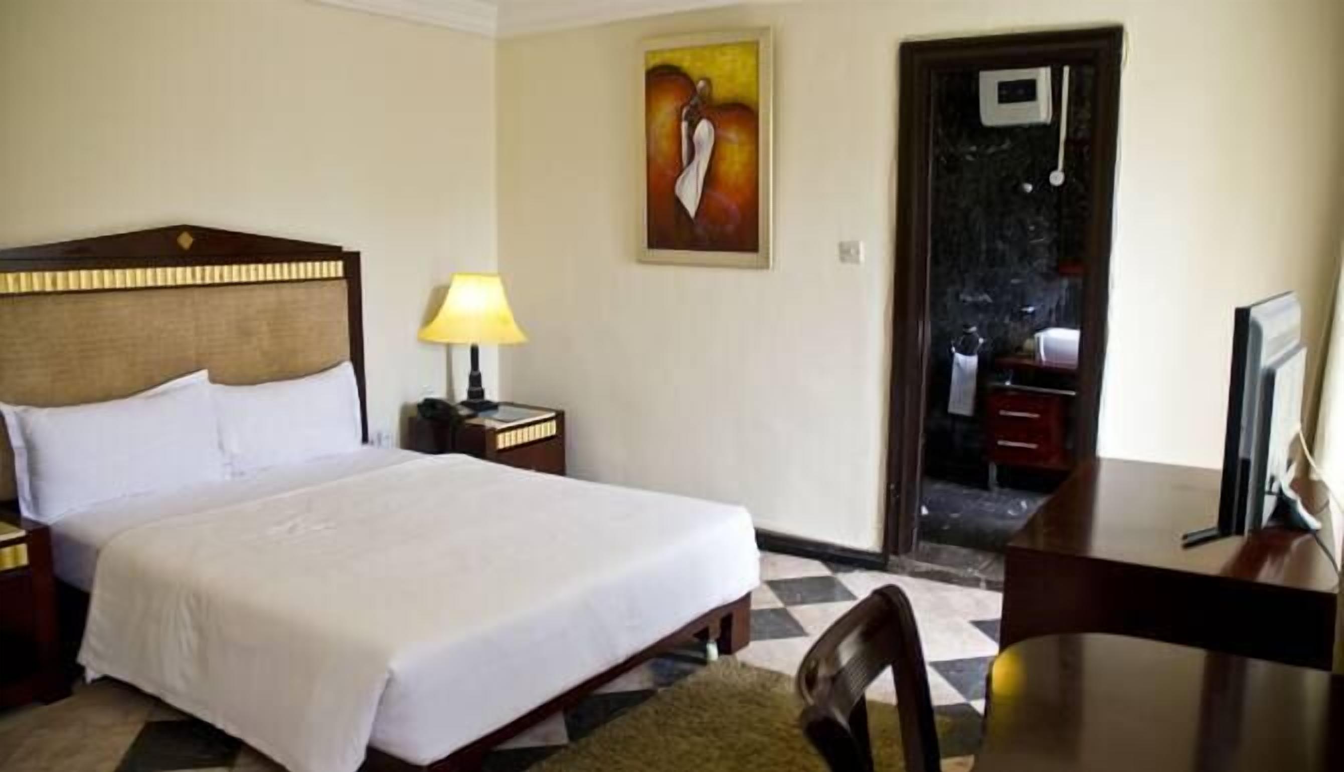 A hotel room with a bed, a desk, a chair, a lamp, and a painting on the wall.