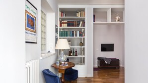 A room with a bookshelf, a table with a lamp, a blue chair, a wooden floor, and a TV on the wall. - Celestina House - Prati, Rome (updated 2026) (Roma)