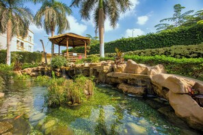 Un estanque tranquilo con rocas y plantas, un pabellón de madera y un edificio al fondo.