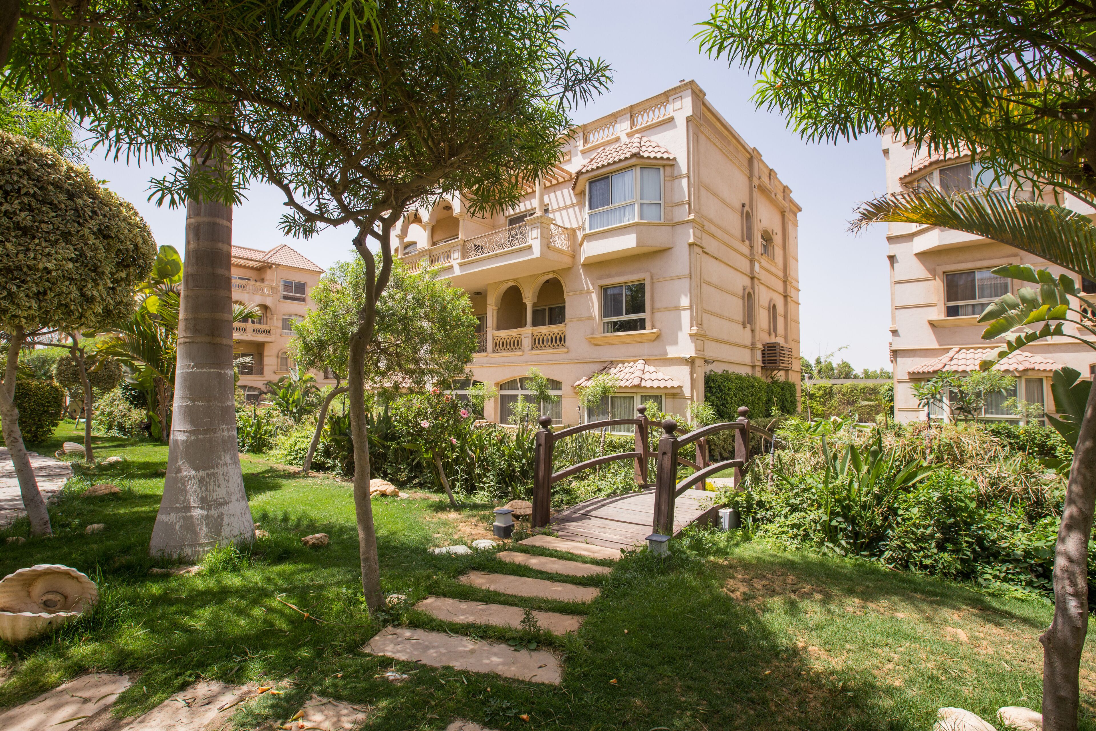Um hotel com vários andares e um jardim com uma ponte de madeira, degraus de pedra e muita vegetação.