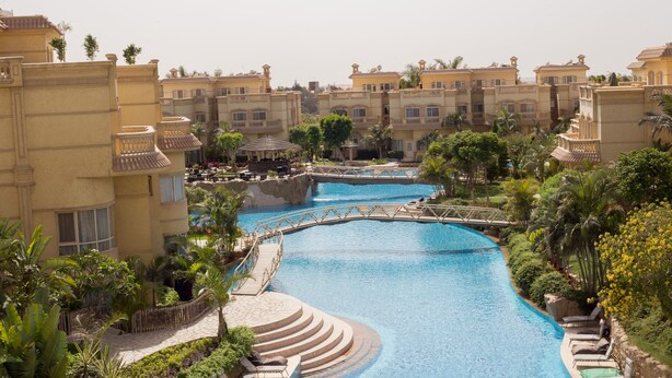 Un resort con una piscina central conectada por un puente, rodeado de exuberante vegetación y varios edificios con balcones.