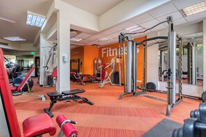 Un gimnasio con diversos equipos de ejercicio, incluyendo máquinas de pesas y pesas libres, con un espejo grande en la pared y suelo alfombrado.