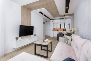 A modern living room with a flat-screen TV, a sofa, a coffee table, and a kitchen in the background. - Cozy Apartment Balcony Close to the Beach (Kołobrzeg)