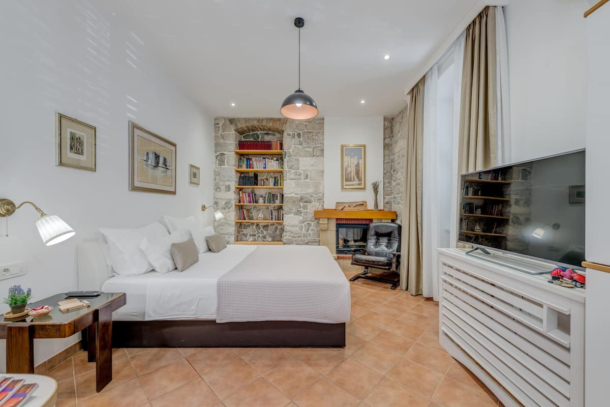 Un dormitorio con cama, mesita de noche, lámpara, estantería, chimenea, televisor y un amplio ventanal con cortinas.