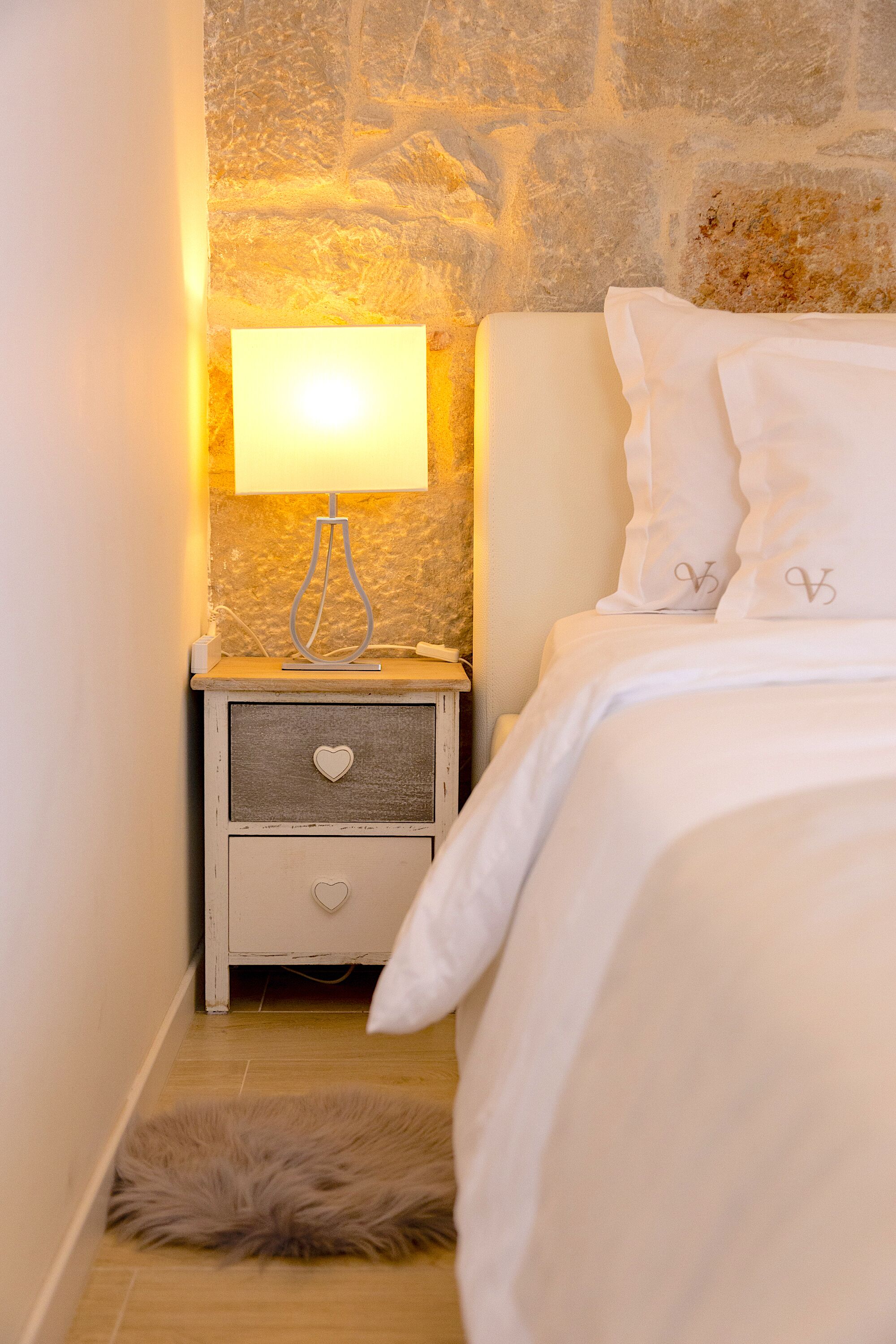 A bedroom with a stone wall, a bedside lamp, a white bed with heart-shaped pillows, and a fluffy rug on the floor.
