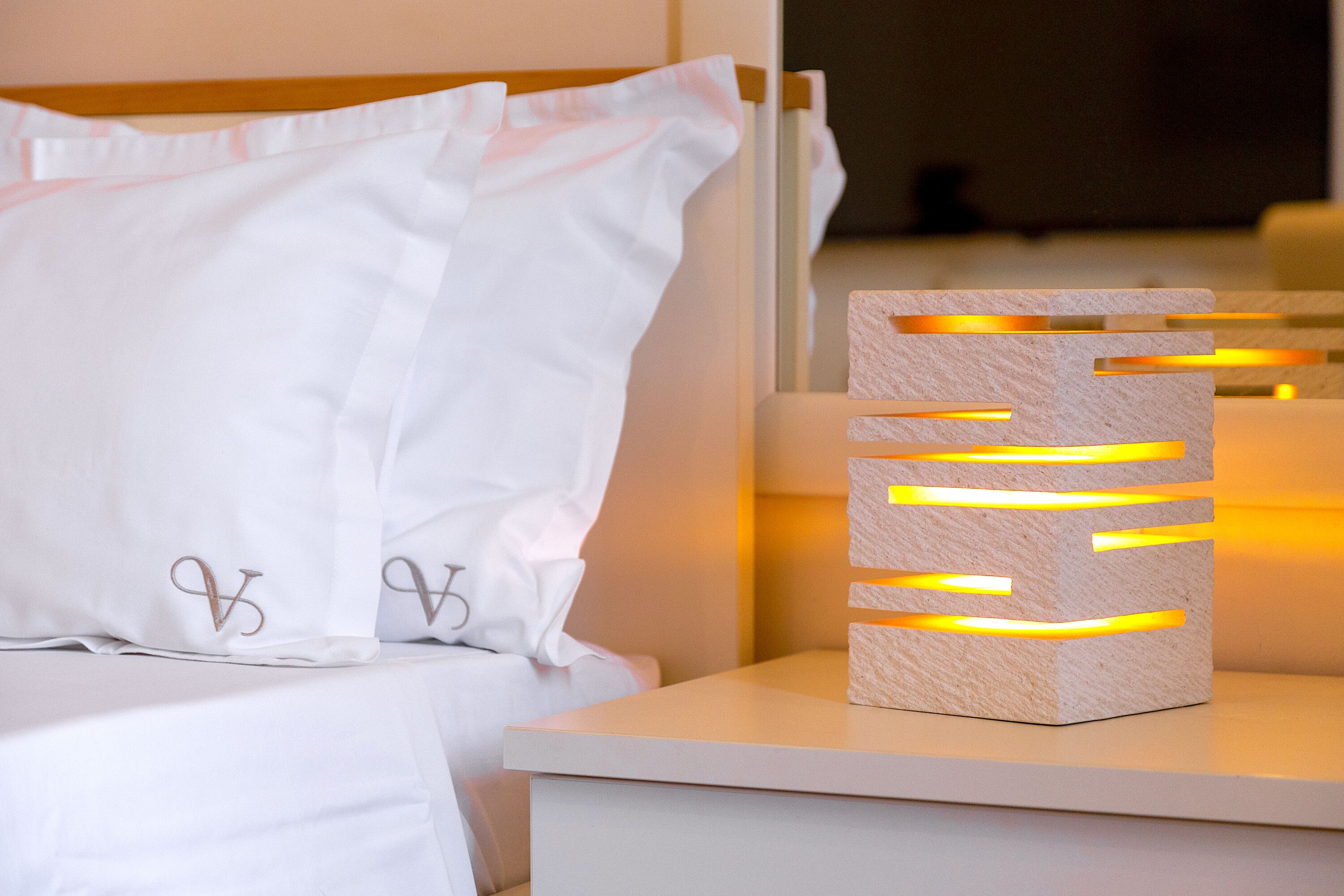 A neatly made bed with white linens and pillows, a bedside table with a modern light fixture, and a wooden headboard.