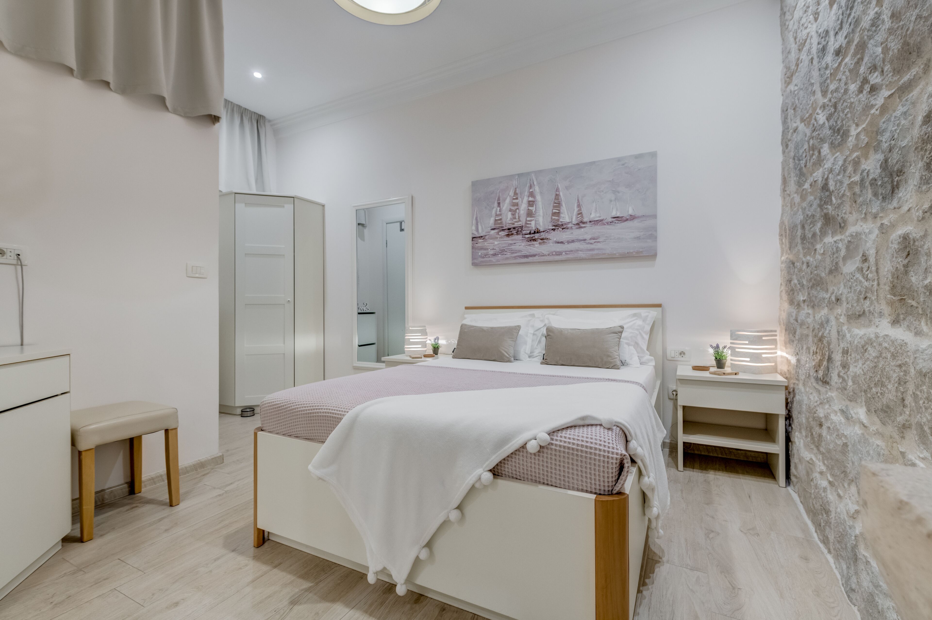 A bedroom with a bed, bedside tables, a bench, a wardrobe, a painting, and a stone wall.