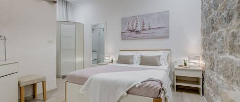 A bedroom with a bed, bedside tables, a bench, a wardrobe, a painting, and a stone wall.