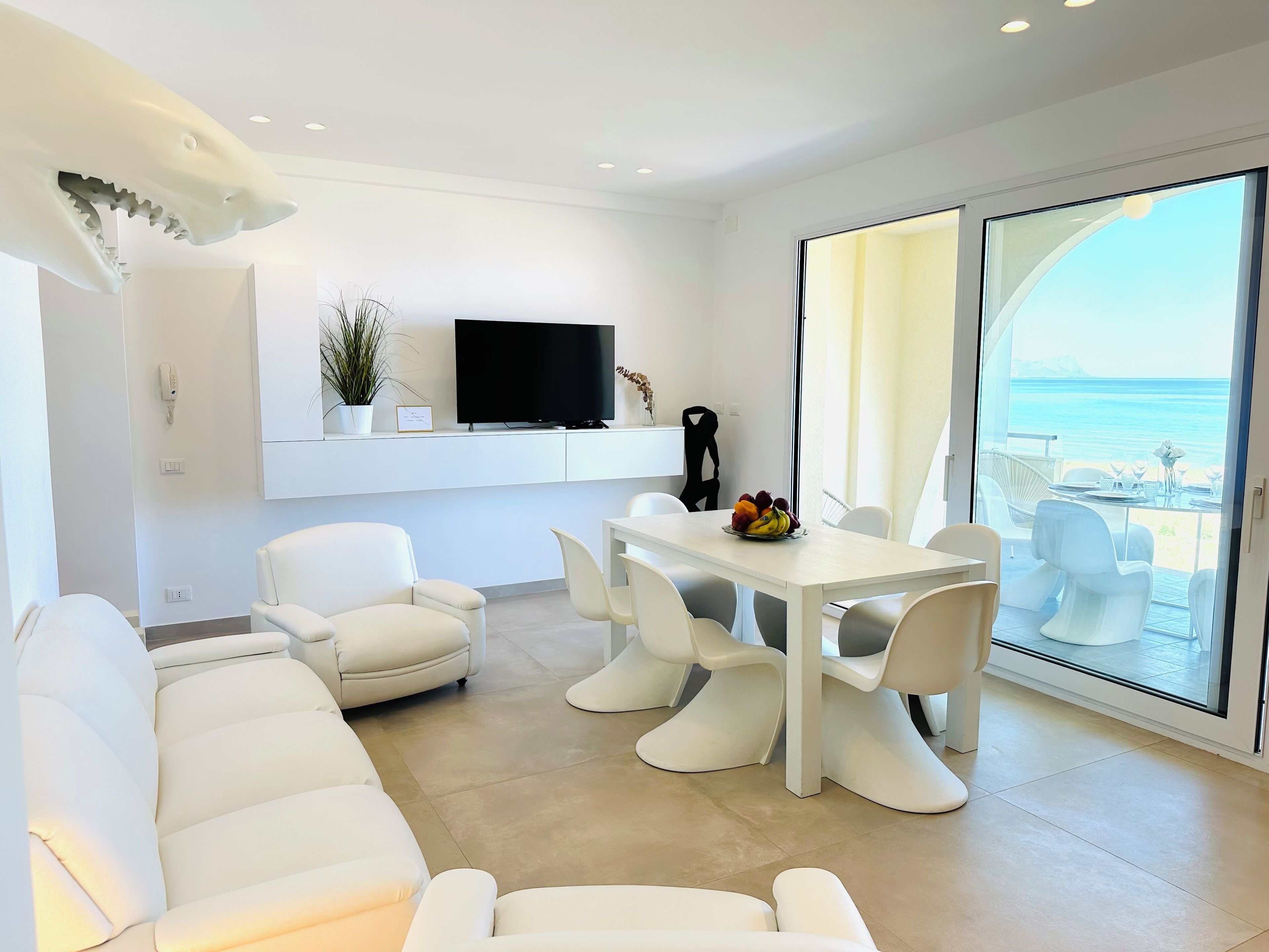 Una sala de estar moderna con un sofá blanco, una mesa de comedor con sillas, un televisor de pantalla plana montado en la pared y un ventanal con vistas al mar.