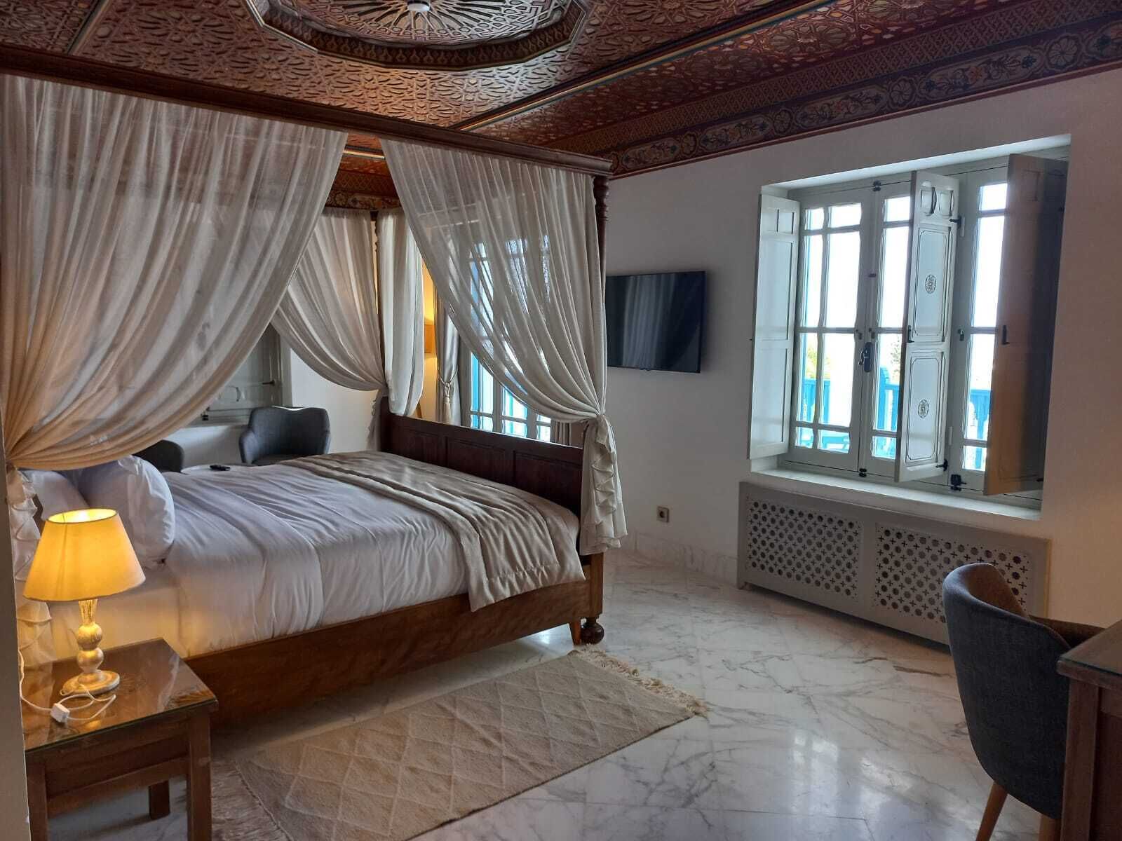 A bedroom with a four-poster bed, a bedside lamp, a window with curtains, and a radiator.