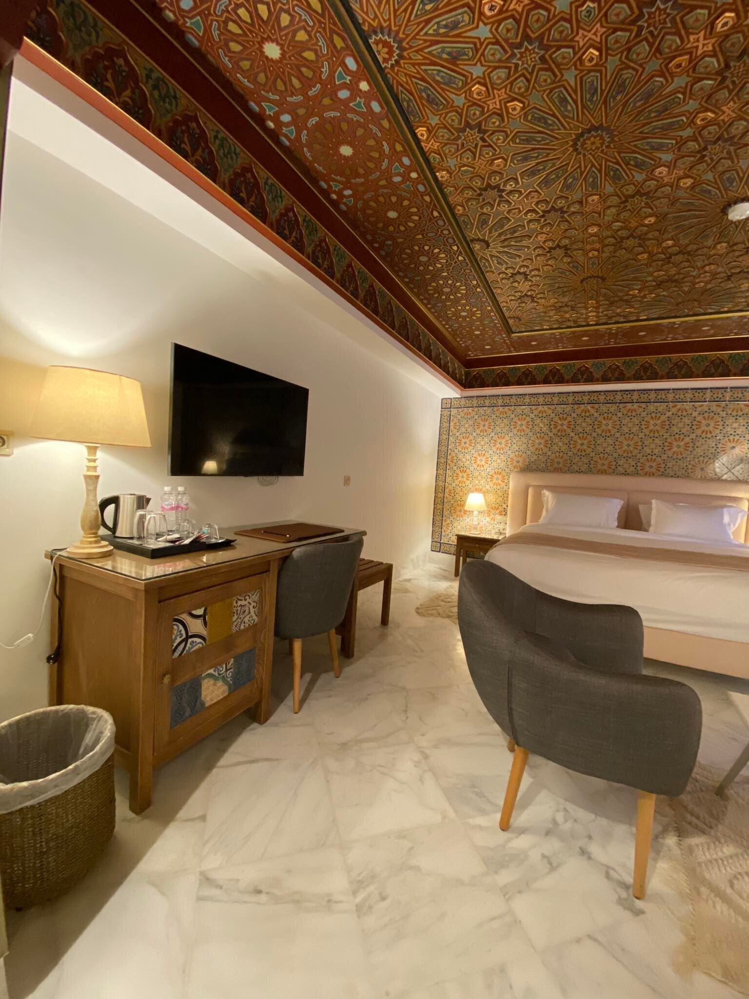 A hotel room with a bed, a desk with a chair, a TV mounted on the wall, and a patterned ceiling.