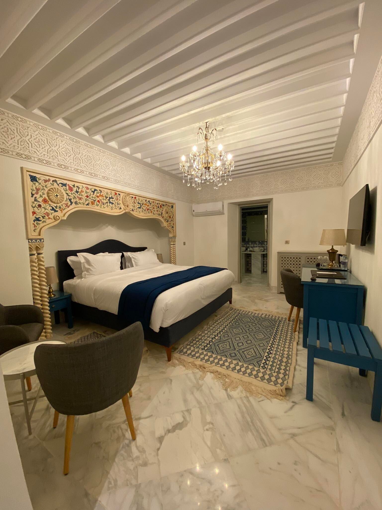 A hotel room with a large bed, a desk, a chair, a TV, and a chandelier.