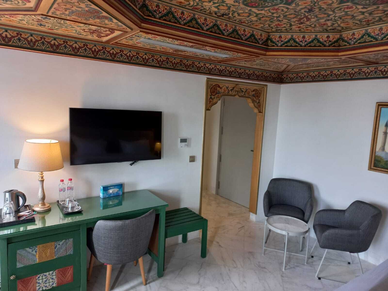 Una camera d'albergo con una scrivania verde, due sedie, una televisione, una lampada e un soffitto decorato.