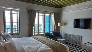 Habitación de hotel con cama, dos sillones, una mesita, televisor y balcón con barandilla azul.