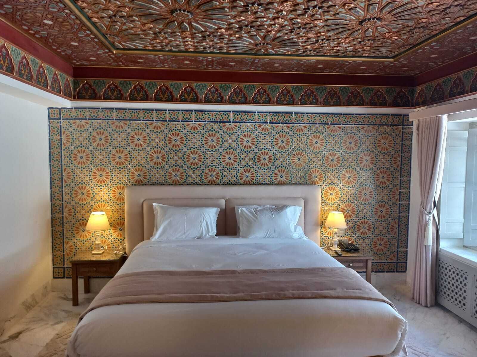 Une chambre d’hôtel avec un grand lit, deux tables de chevet avec des lampes, un papier peint à motifs et un plafond décoratif.