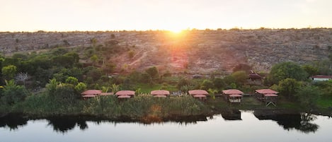 A lakeside view with several small buildings with red roofs, surrounded by greenery and a distant hill.