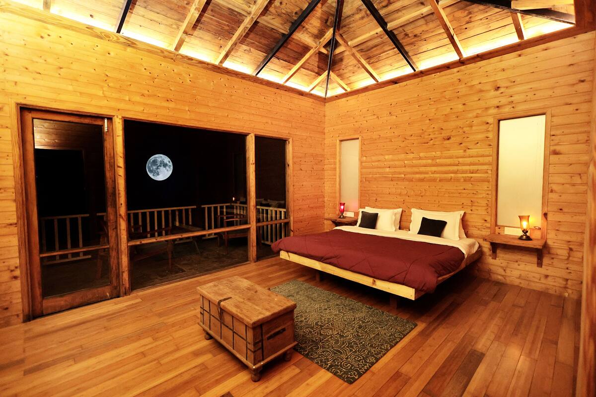 A wooden cabin bedroom with a bed, a nightstand, a bench, and a view of the moon through a large window.