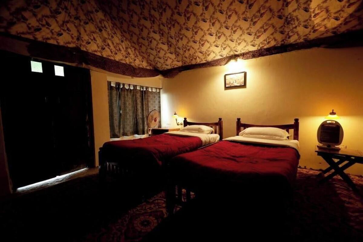 A room with two beds, a patterned ceiling, a small table with a lamp, and a door.