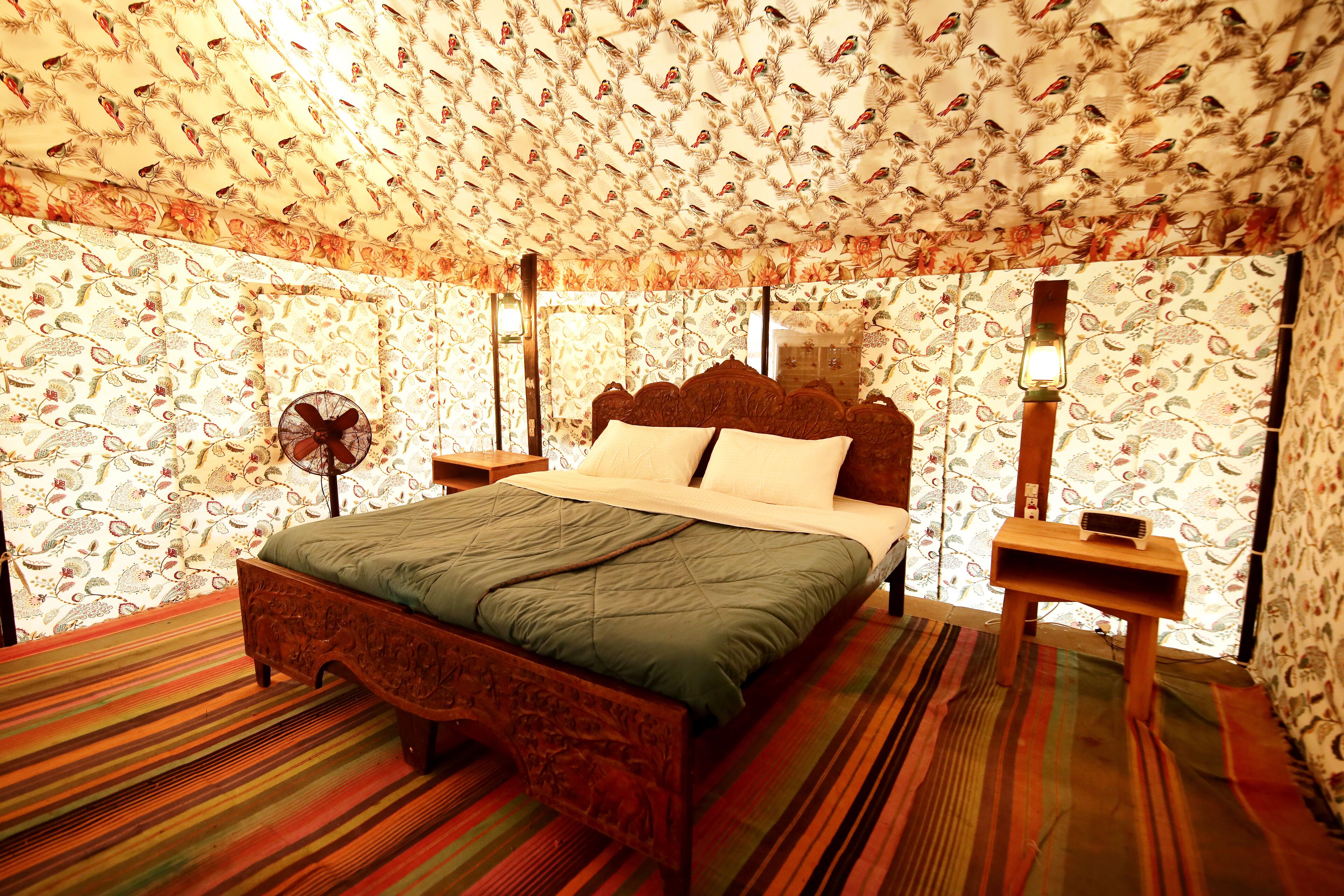 A room with a patterned wallpaper, a bed with a wooden headboard, a bedside table with a lamp, and a striped carpet.