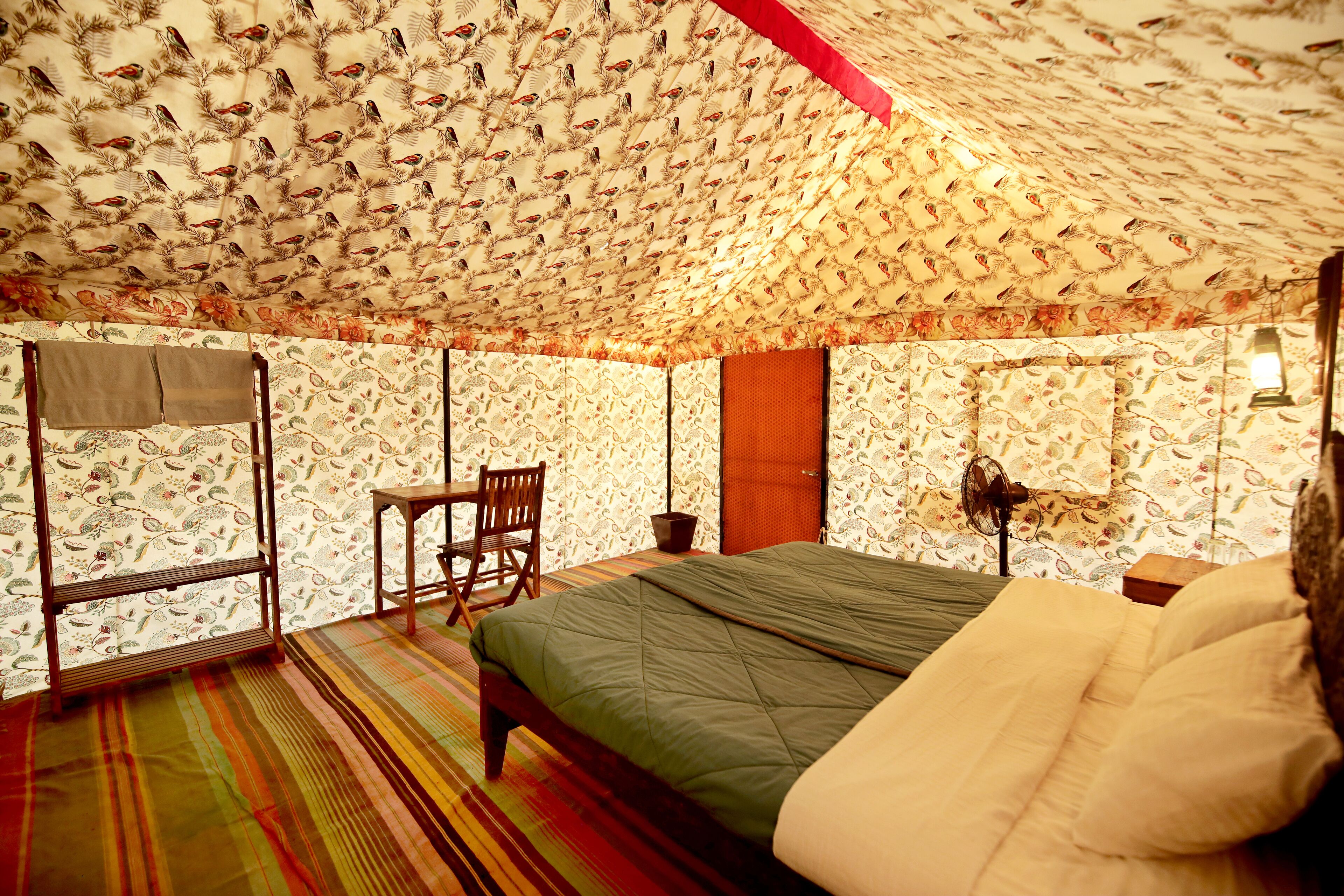 A room with a patterned ceiling, a bed with a green bedspread, a wooden chair, a small table, and a wall-mounted lamp.