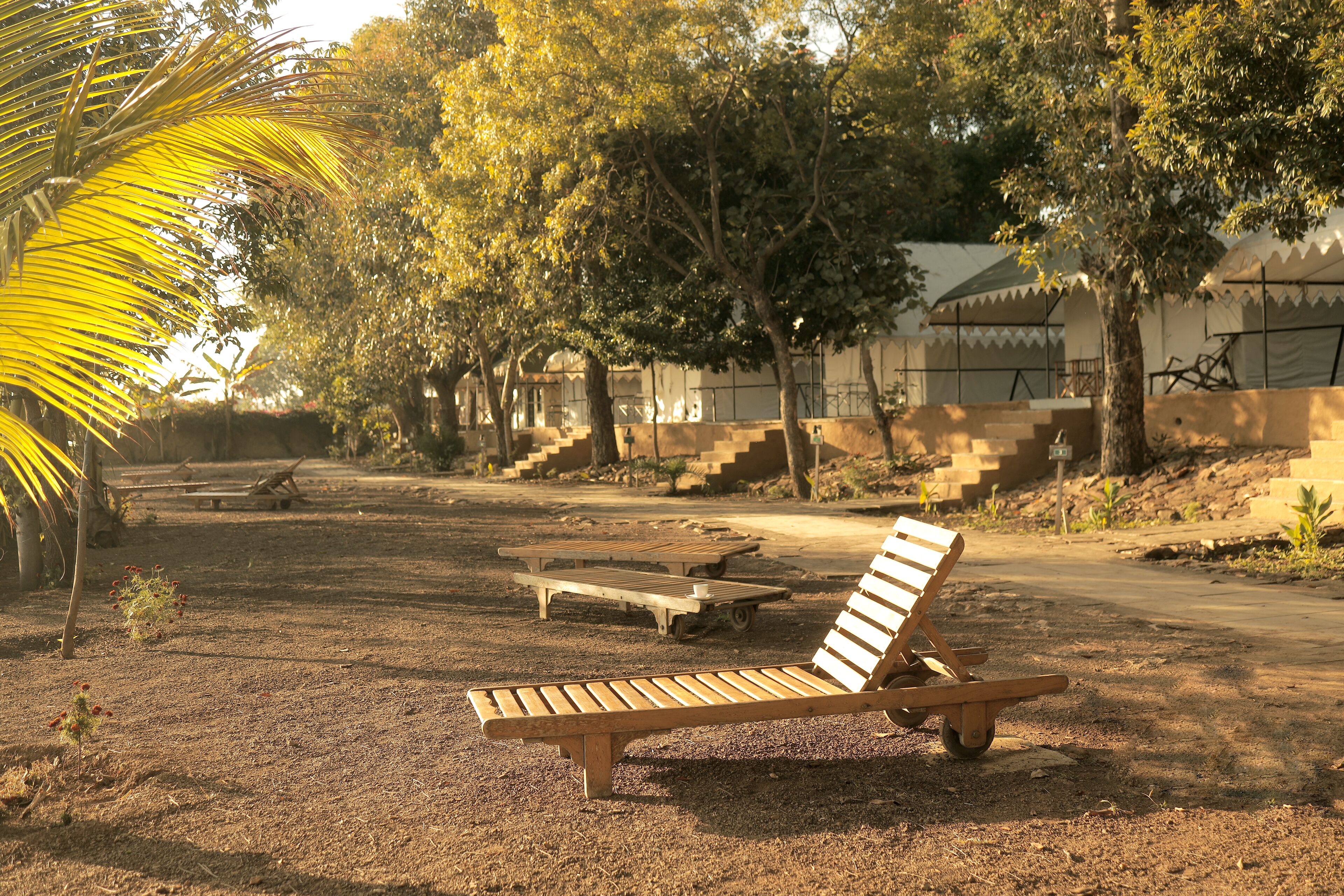 A wooden lounge chair in a shaded outdoor area with a dirt ground, surrounded by trees and a building with a tiled roof in the background.