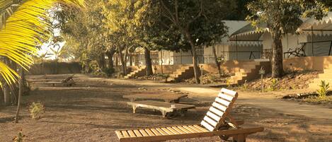 A wooden lounge chair in a shaded outdoor area with a dirt ground, surrounded by trees and a building with a tiled roof in the background.