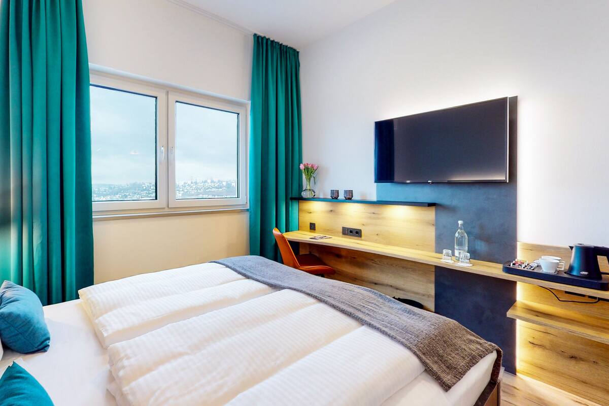 Habitación de hotel con cama, escritorio, silla, televisión y ventana con cortinas.