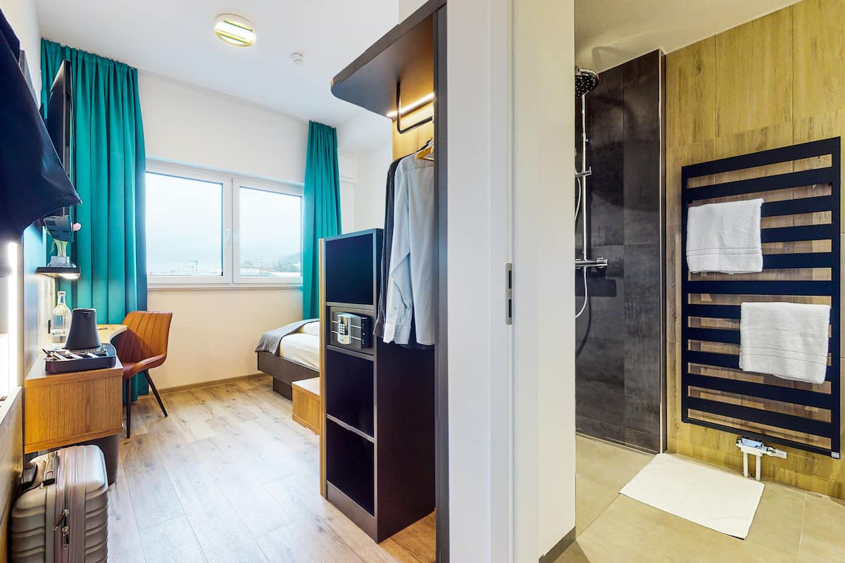 Habitación de hotel con cama, escritorio con lámpara, televisor, armario, zona de ducha y ventana con cortinas.