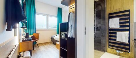 A hotel room with a bed, a desk with a lamp, a TV, a wardrobe, a shower area, and a window with curtains.