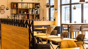 A modern cafe with wooden tables and black chairs, hanging pendant lights, and a bookshelf in the background. - Luma Hotel Spa&Fitness (Fulda)