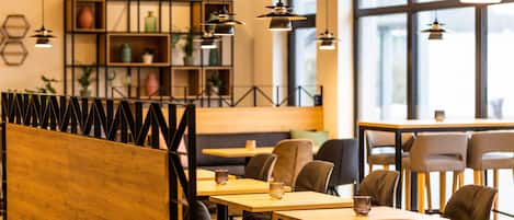 Ein modernes Café mit Holztischen und schwarzen Stühlen, hängenden Pendelleuchten und einem Bücherregal im Hintergrund.