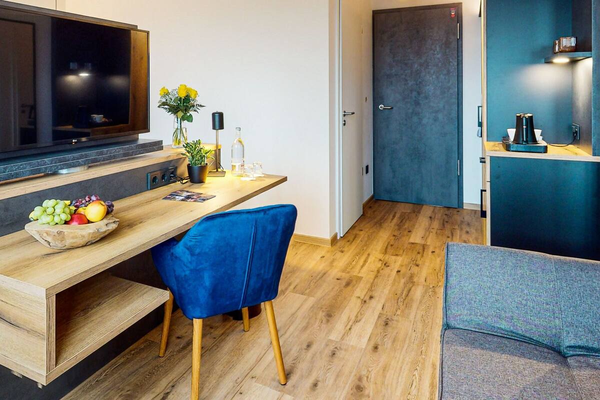 Una habitación de hotel moderna con una cama, un escritorio con computadora, televisor y una zona de estar con sofá y sillón.