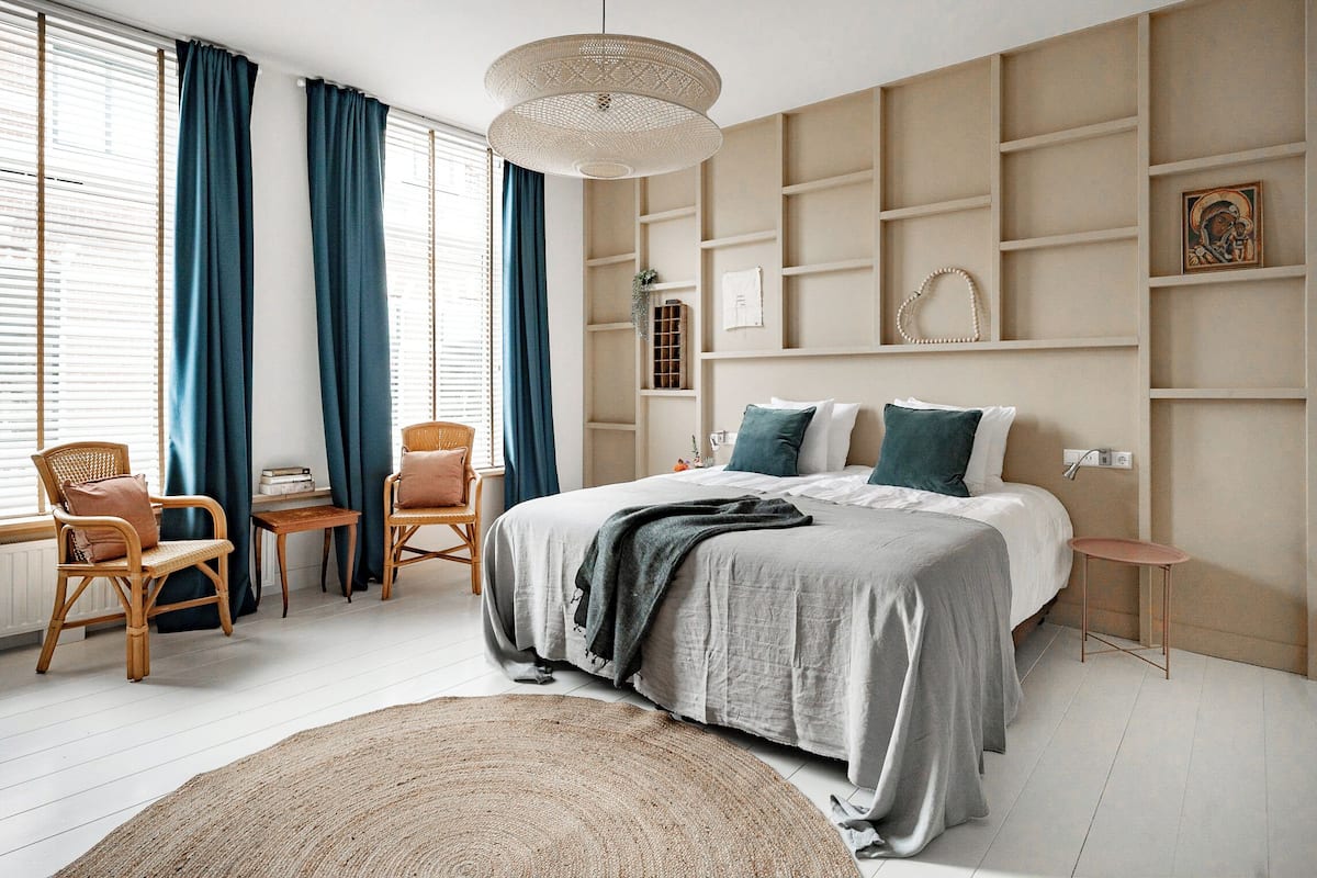 A bedroom with a large bed, two chairs, a small table, a hanging light fixture, and built-in shelving.
