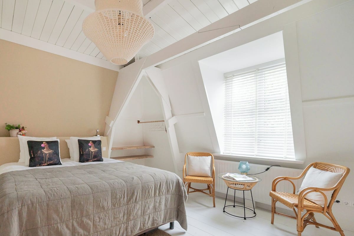 A bedroom with a large bed, two chairs, a small table, and a hanging light fixture.