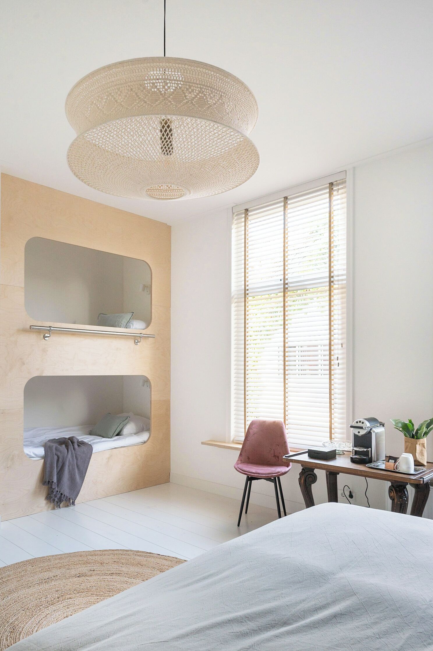 A modern bedroom with a bunk bed, a desk with a chair, a large window with blinds, and a woven pendant light.