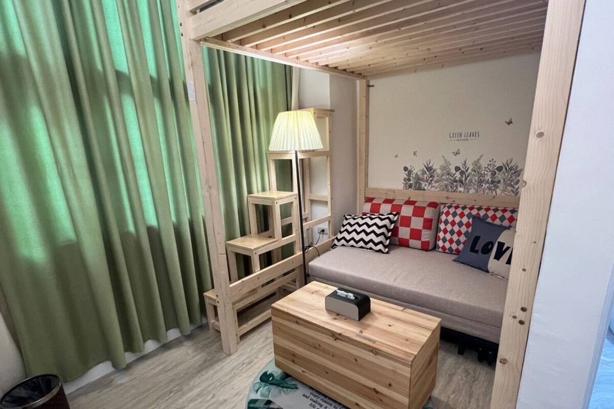 A cozy room with a wooden bed, a small wooden table, a lamp, and a sofa with patterned cushions.