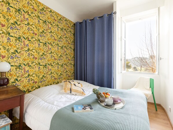 Un dormitorio con papel tapiz floral, una cama con edredón blanco, una cortina azul, un ventanal con vistas, una mesita de noche de madera, una lámpara, un sillón verde y una bandeja con frutas y tazas.