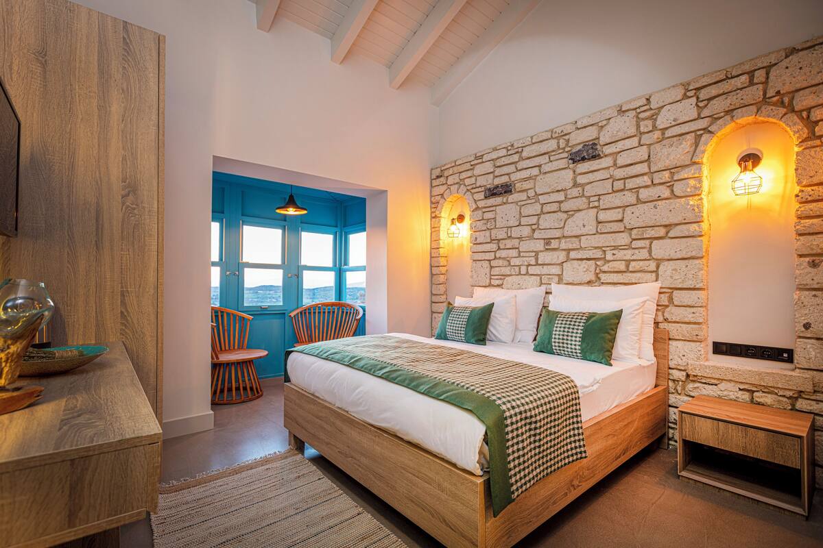Un dormitorio con pared de piedra, cama con ropa de cama verde y blanca, mesita de noche de madera y vistas a través de una puerta azul.