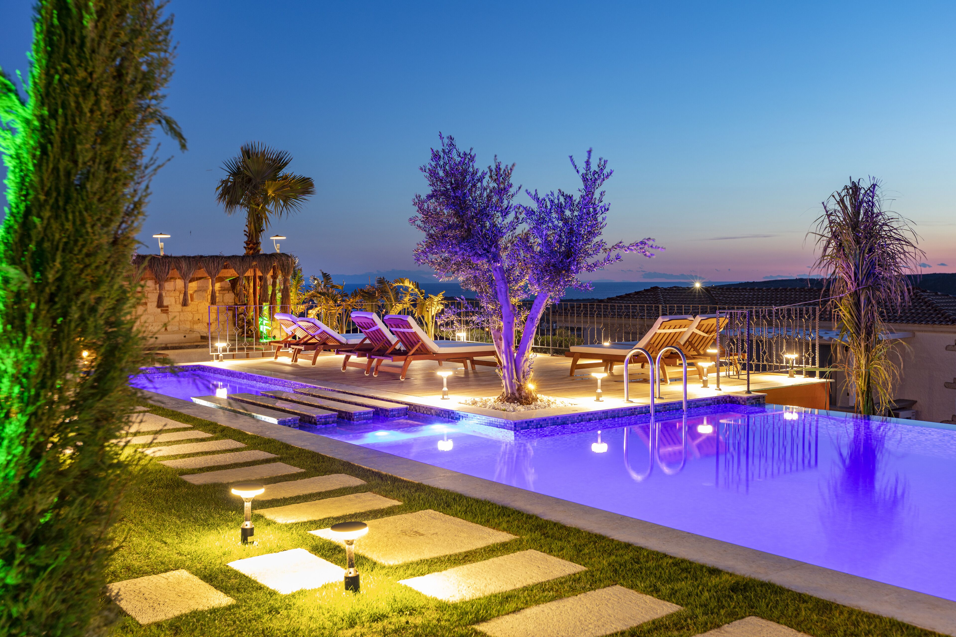 A swimming pool with lounge chairs and a tree, surrounded by a well-lit garden area.