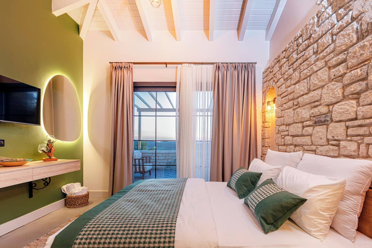 Un dormitorio con una cama grande, televisor, pared de piedra y balcón con cortinas.