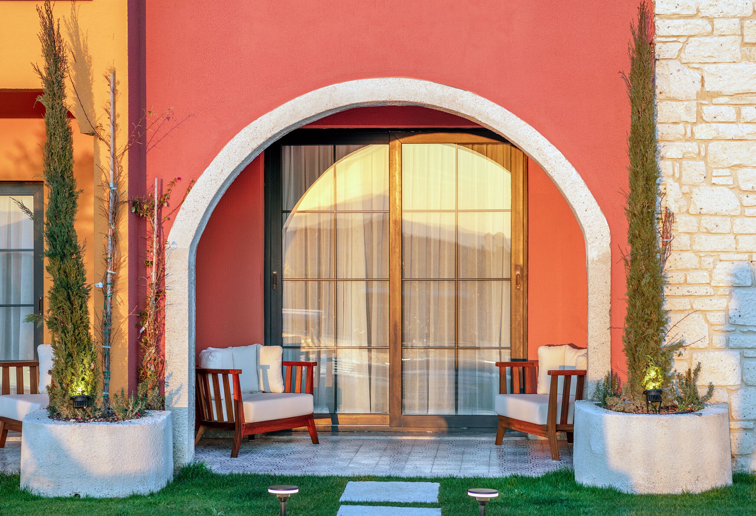 Un bâtiment doté d’une grande entrée en arc de cercle, de deux espaces de détente avec des chaises et des coussins, et de plantes en pot de chaque côté.