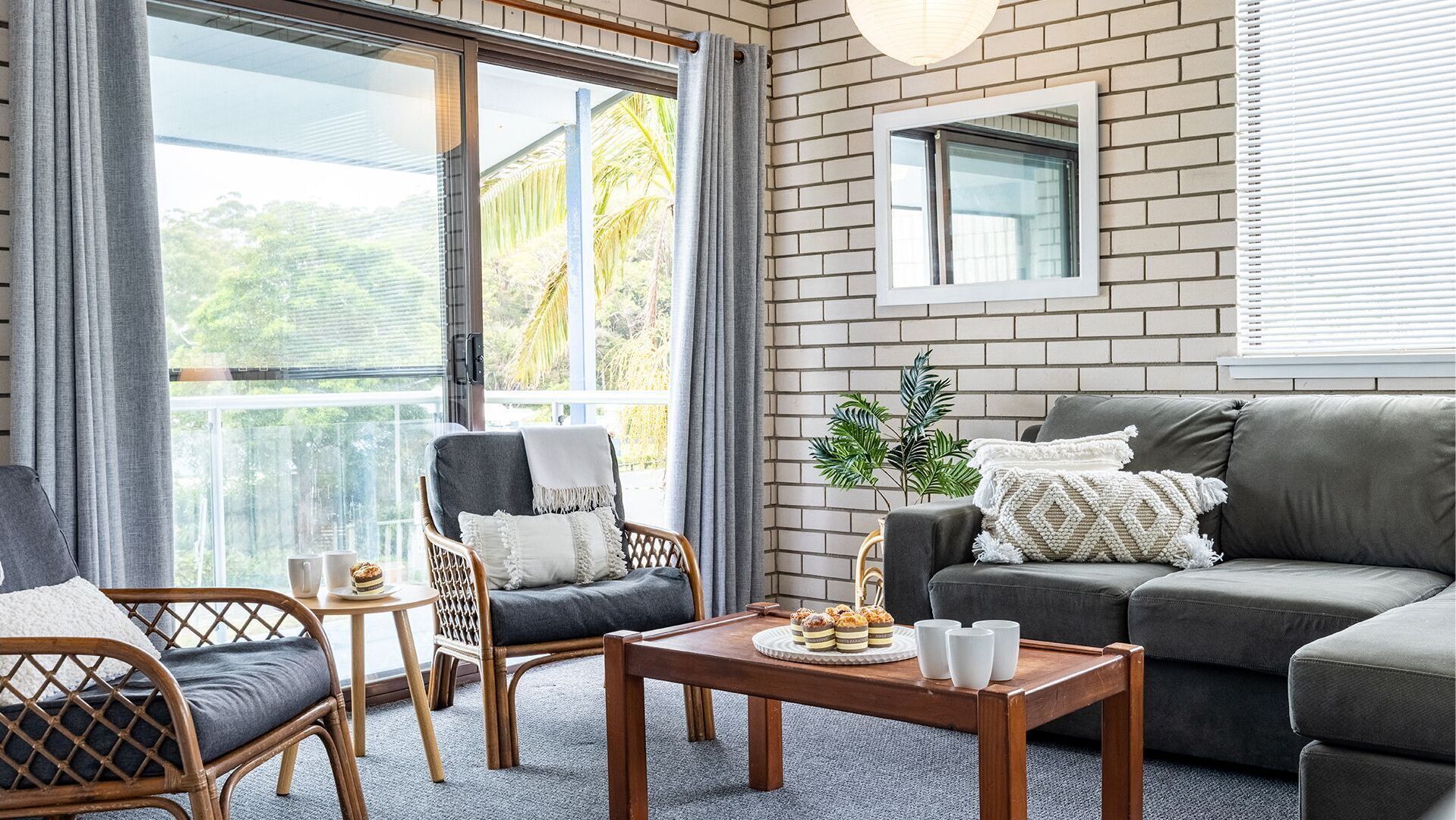 A modern living room with a grey sofa, wicker chairs, a wooden coffee table, and a balcony with a view of greenery.