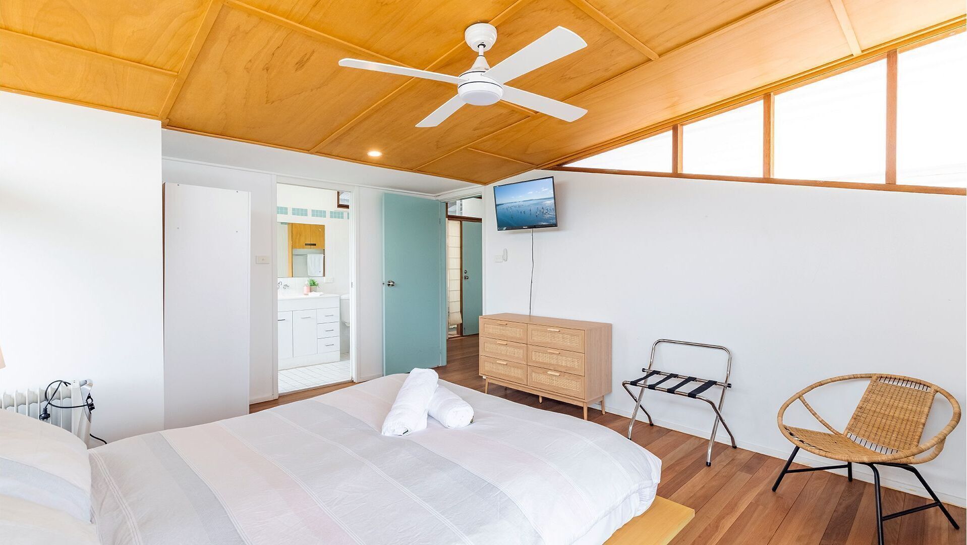 A bedroom with a bed, a ceiling fan, a TV, a dresser, a chair, and a bathroom visible through an open door.