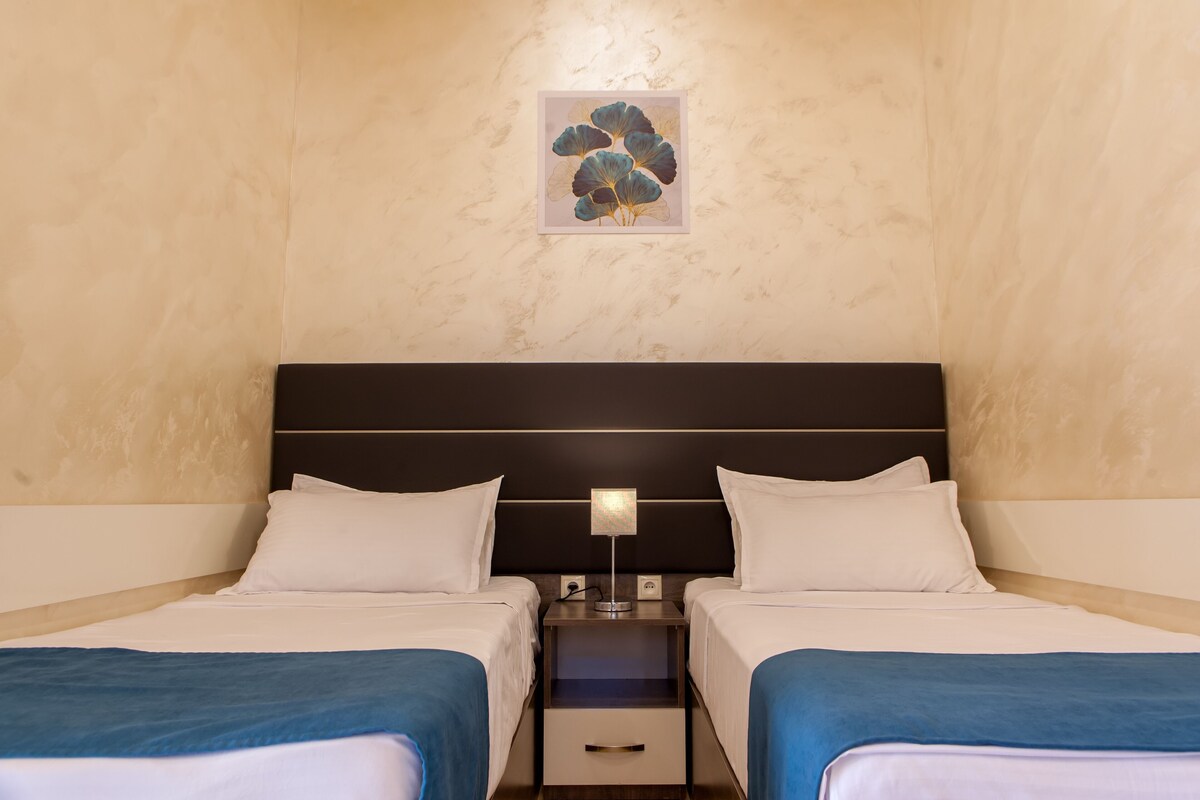 A hotel room with two beds, a nightstand with a lamp, a small drawer, and a wall-mounted artwork.