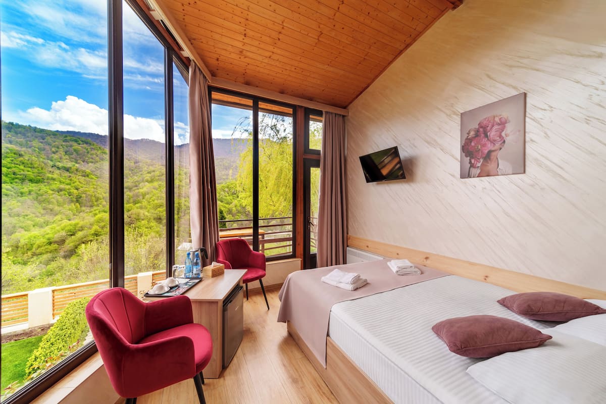 A bedroom with a large window, a desk, a chair, and a bed with pillows.