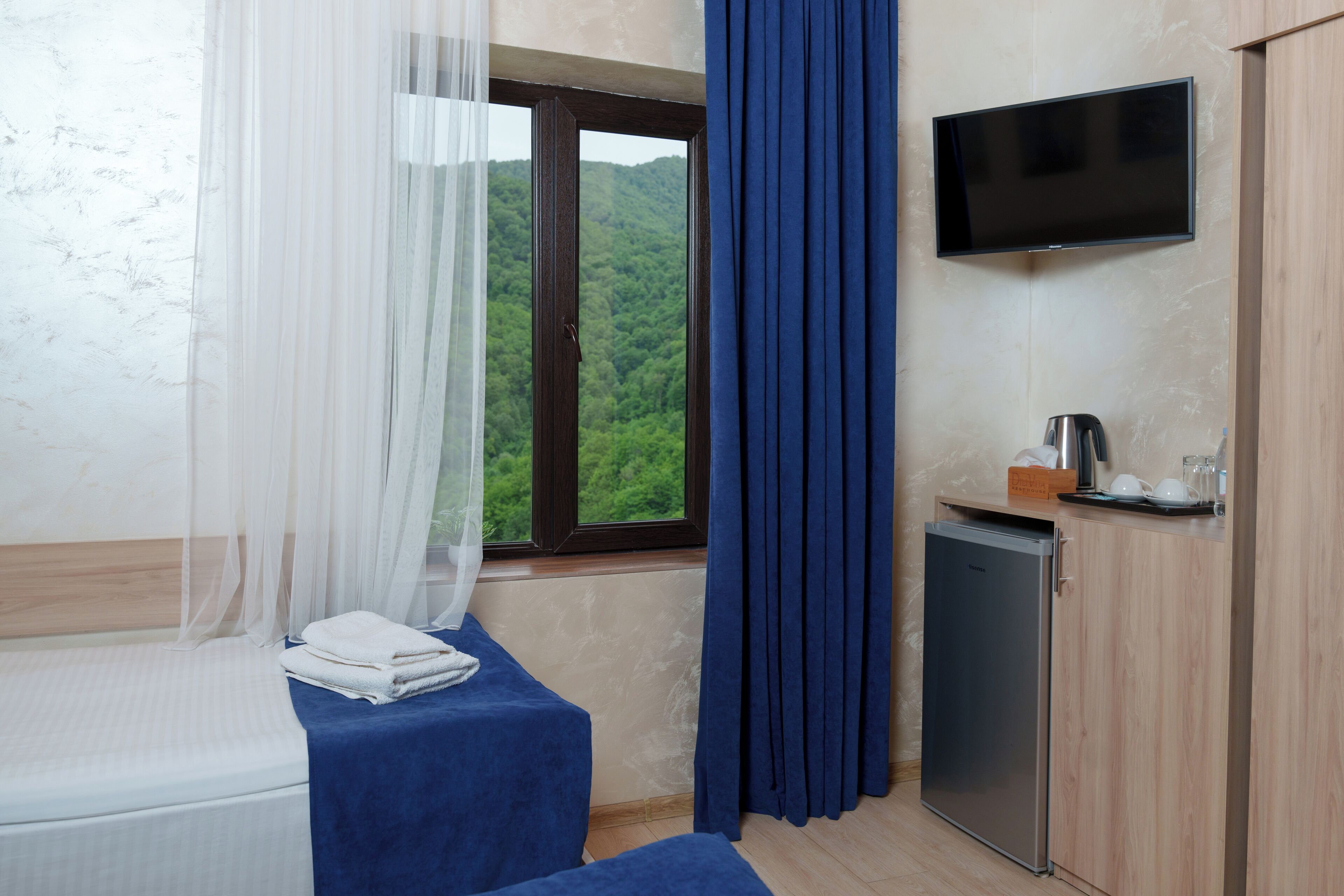 Un dormitorio con cama, una mesita con hervidor, televisión y vista a un bosque por la ventana.